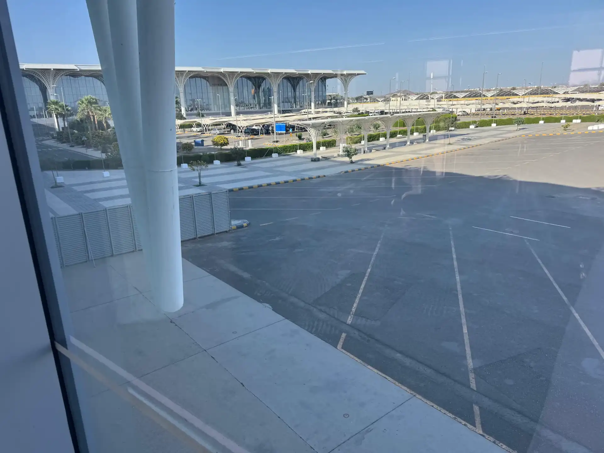 View through the terminal glass of the Madinah Airport facade with arched columns, palm trees and the car park.